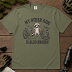 Moss green t-shirt with raccoon graphic and text on a wooden surface with outdoor gear.