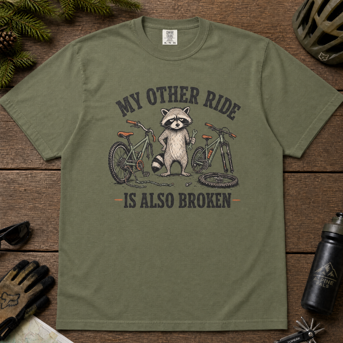 Moss green t-shirt with raccoon graphic and text on a wooden surface with outdoor gear.