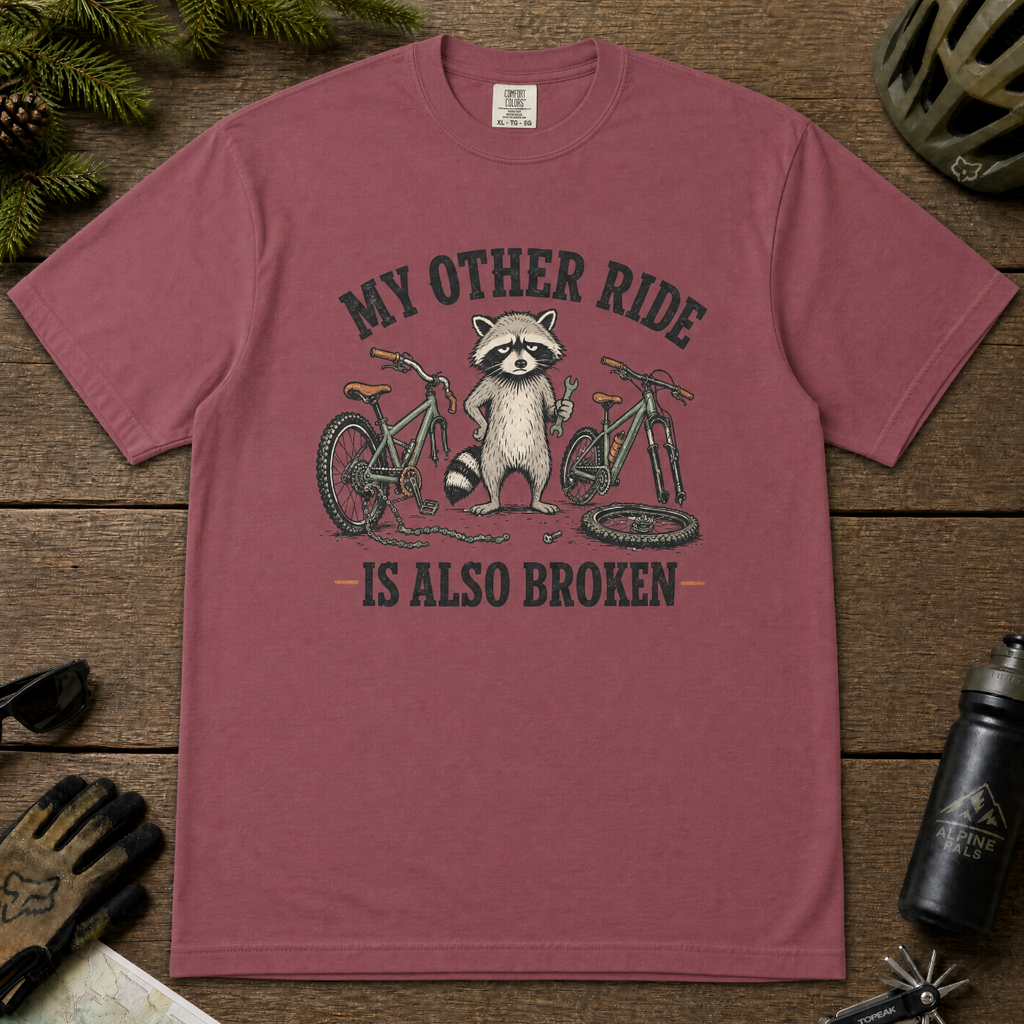 Brick red t-shirt with a raccoon and bicycle graphic on a wooden surface with outdoor gear.