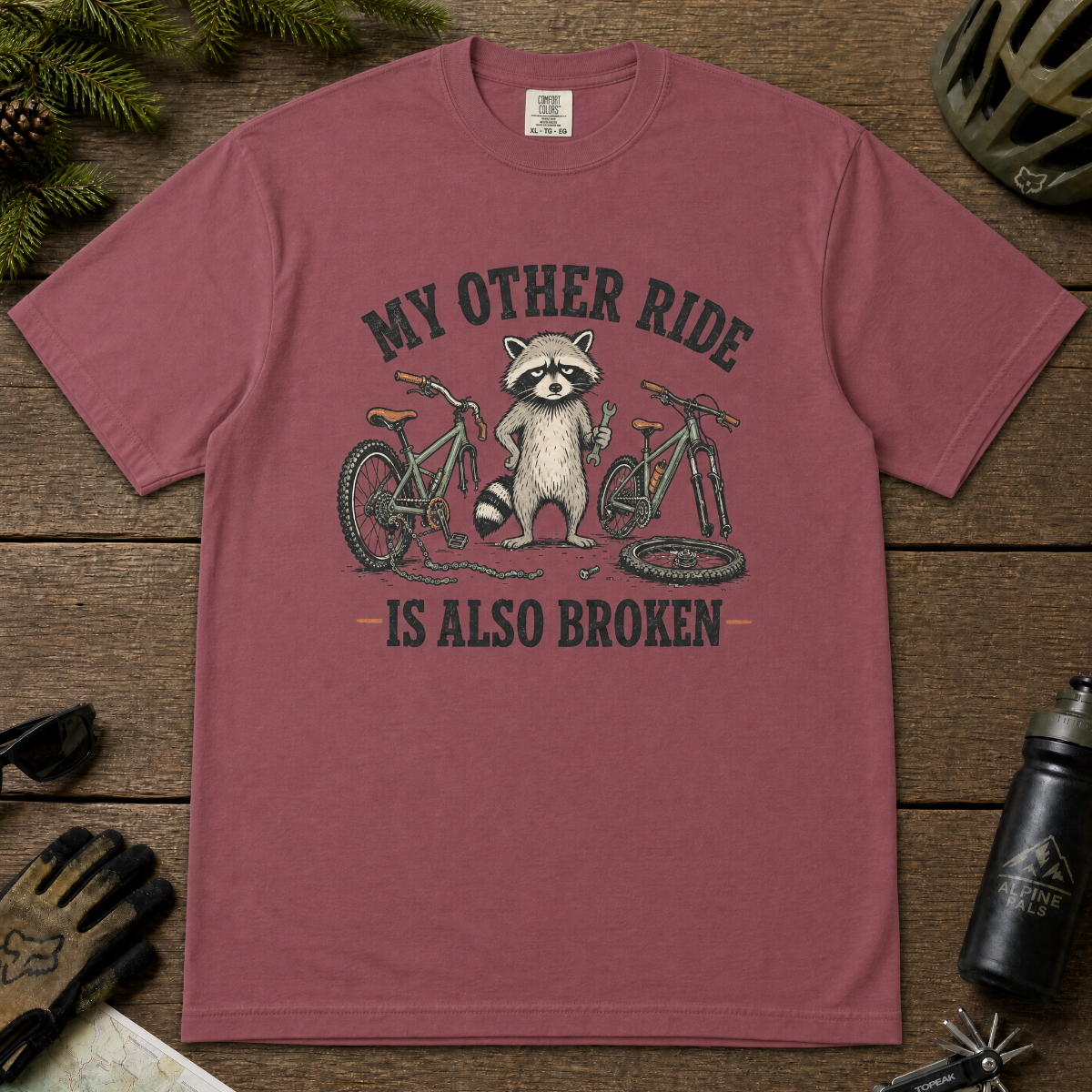 Brick red t-shirt with a raccoon and bicycle graphic on a wooden surface with outdoor gear.