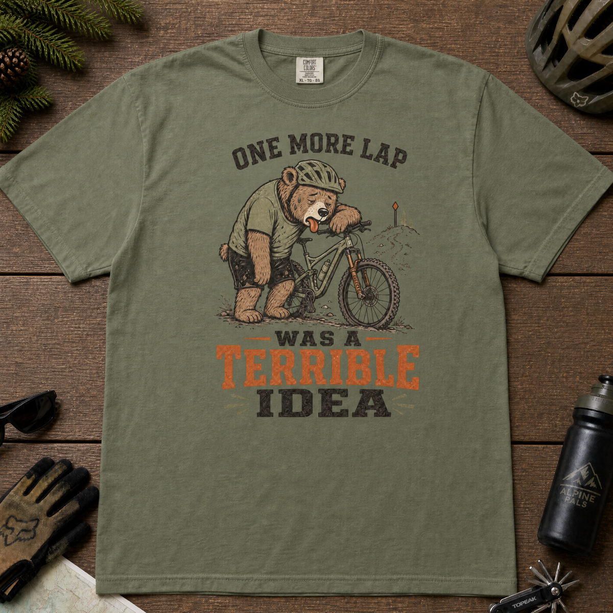 Moss green t-shirt with bear graphic and text on a wooden surface with outdoor gear.