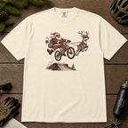 Ivory white t-shirt with a graphic of Santa Claus riding a bike with a reindeer, placed on a wooden surface with outdoor gear.