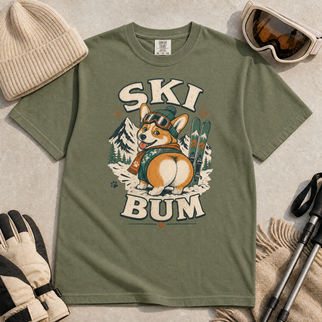 Moss green t-shirt with a corgi graphic and 'SKI BUM' text, surrounded by winter accessories on a light background.