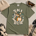 Moss green t-shirt with a corgi graphic and 'SKI BUM' text, surrounded by winter accessories on a light background.
