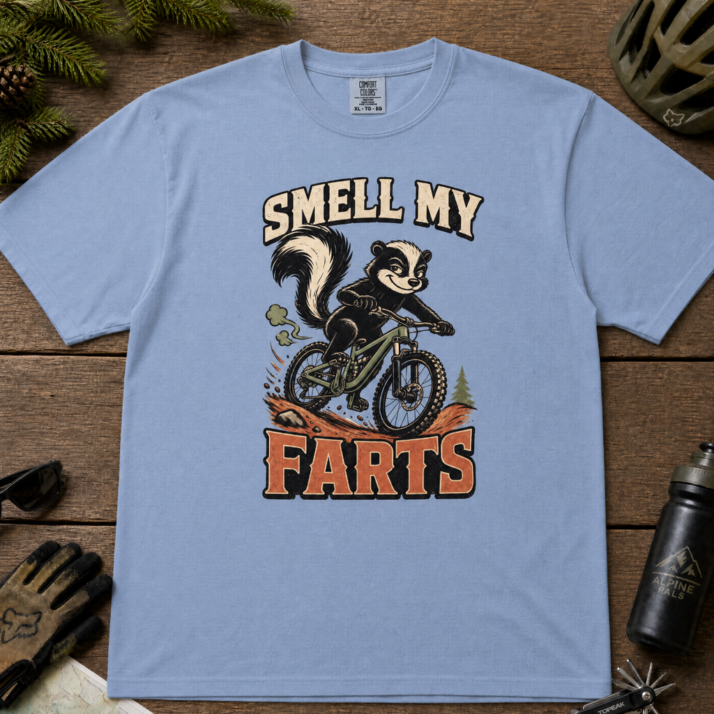 Washed denim blue t-shirt with a skunk on a bike graphic and 'Smell My Farts' text on a wooden surface.