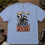Washed denim blue t-shirt with a skunk on a bike graphic and 'Smell My Farts' text on a wooden surface.