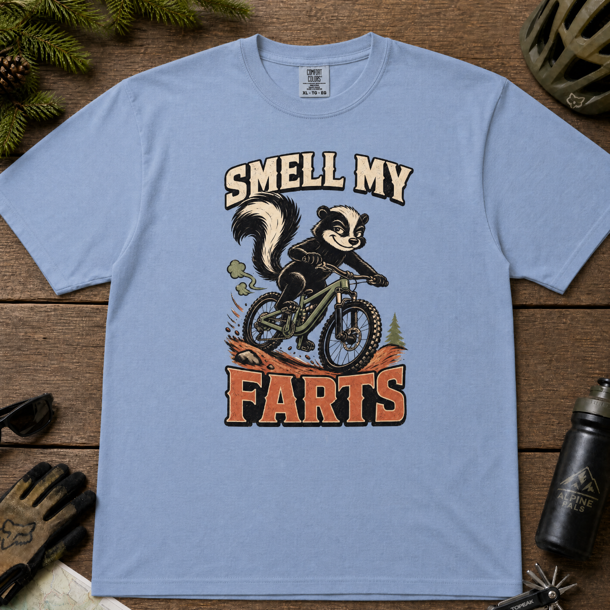 Washed denim blue t-shirt with a skunk on a bike graphic and 'Smell My Farts' text on a wooden surface.