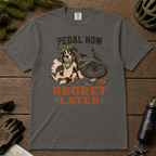 Pepper gray t-shirt with a dog and bicycle graphic and 'Pedal Now Regret Later' text on a wooden surface with cycling accessories.