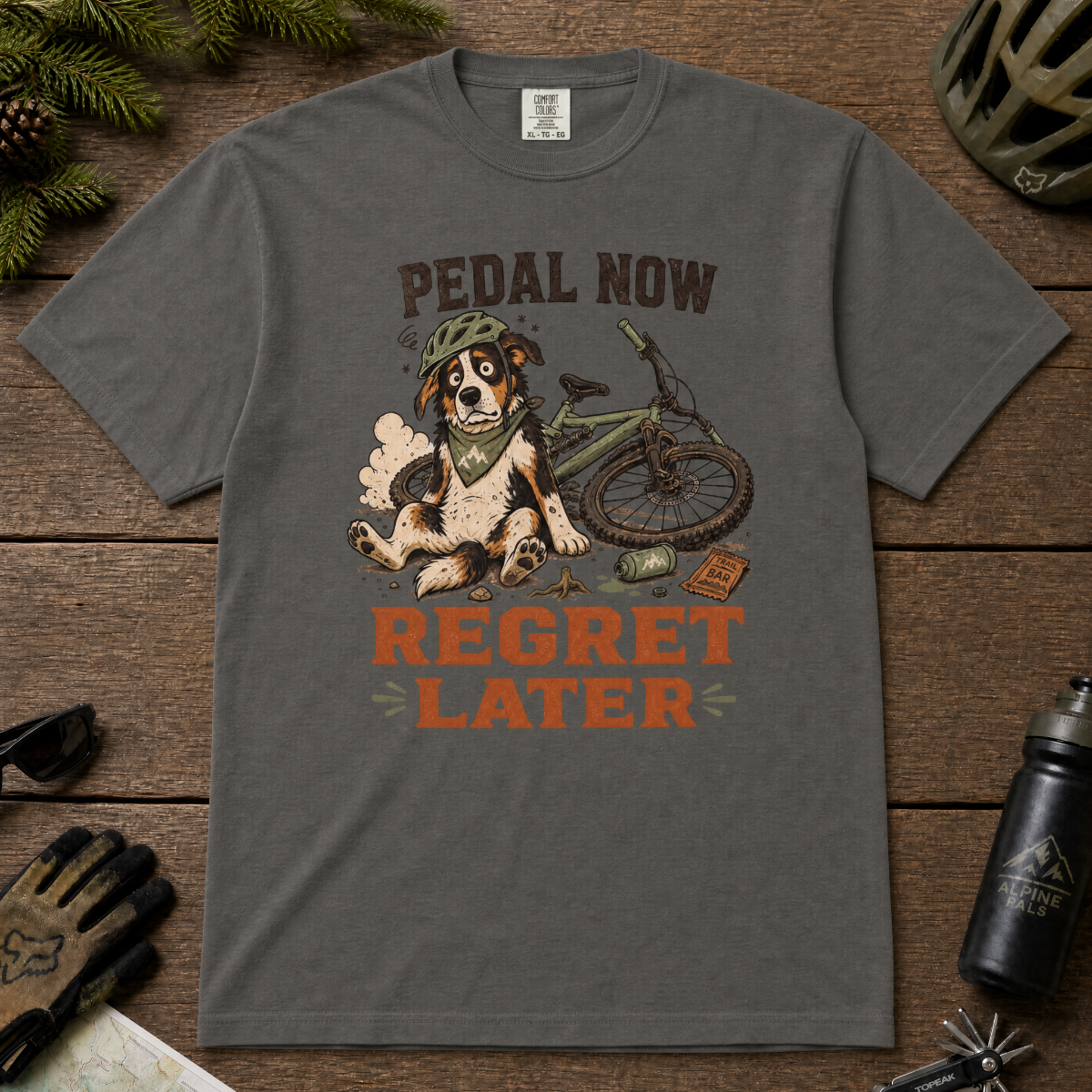 Pepper gray t-shirt with a dog and bicycle graphic and 'Pedal Now Regret Later' text on a wooden surface with cycling accessories.