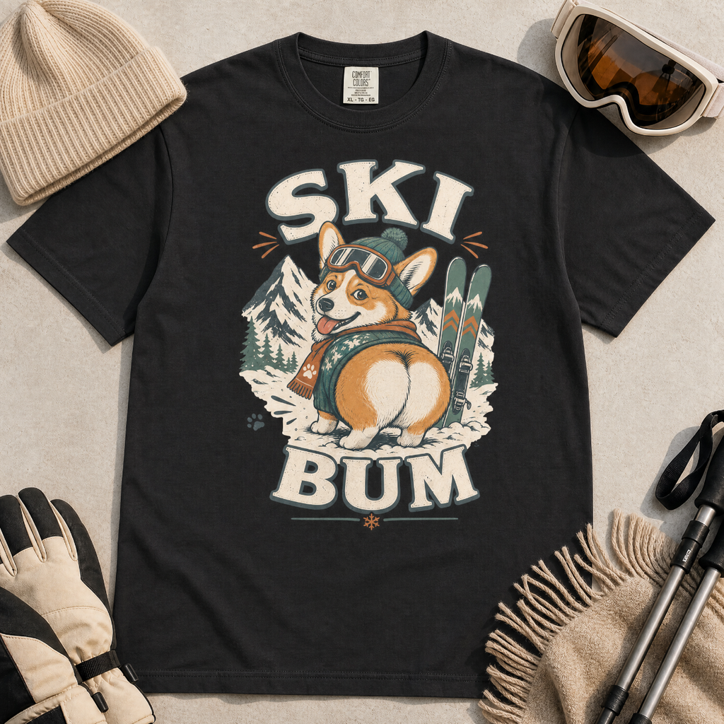 Black t-shirt with a corgi graphic and 'Ski Bum' text, surrounded by winter accessories on a light background.