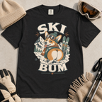 Black t-shirt with a corgi graphic and 'Ski Bum' text, surrounded by winter accessories on a light background.