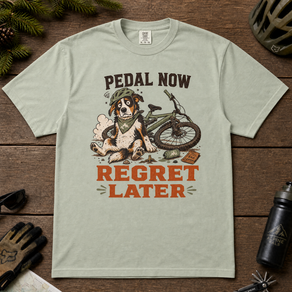 Bay green t-shirt with a dog and bicycle graphic and 'Pedal Now Regret Later' text on a wooden surface.