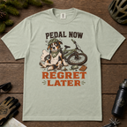 Bay green t-shirt with a dog and bicycle graphic and 'Pedal Now Regret Later' text on a wooden surface.