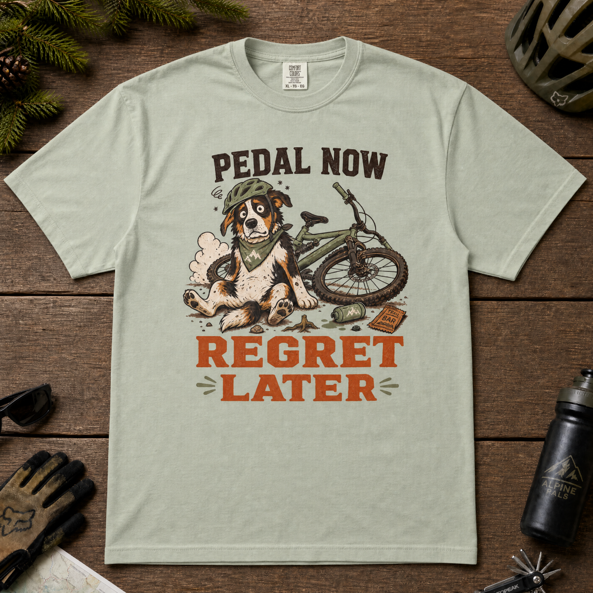 Bay green t-shirt with a dog and bicycle graphic and 'Pedal Now Regret Later' text on a wooden surface.