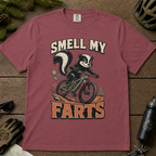 Brick red t-shirt with a graphic of a skunk on a bike and text 'Smell My Farts' on a wooden surface.