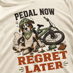 T-shirt with a dog and bicycle graphic and text 'Pedal Now Regret Later' on an ivory white background.