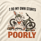 T-shirt with a graphic of a rabbit on a bike and text 'I do my own stunts poorly' on an ivory white background.