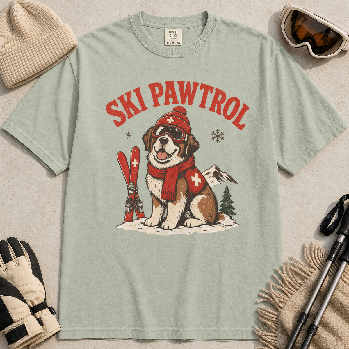 Bay green t-shirt with a dog graphic and 'Ski Pawtrol' text, surrounded by winter accessories on a light gray background.