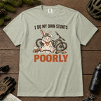 Bay green t-shirt with a graphic of a rabbit and bicycle, text 'I do my own stunts poorly', on a wooden surface with outdoor gear.