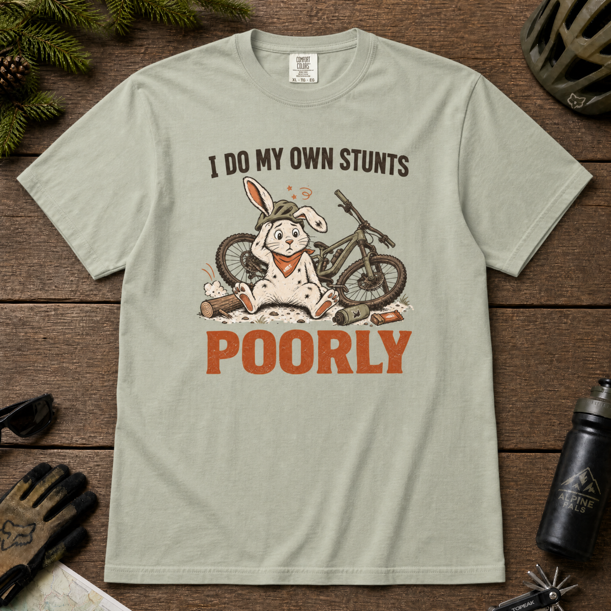 Bay green t-shirt with a graphic of a rabbit and bicycle, text 'I do my own stunts poorly', on a wooden surface with outdoor gear.