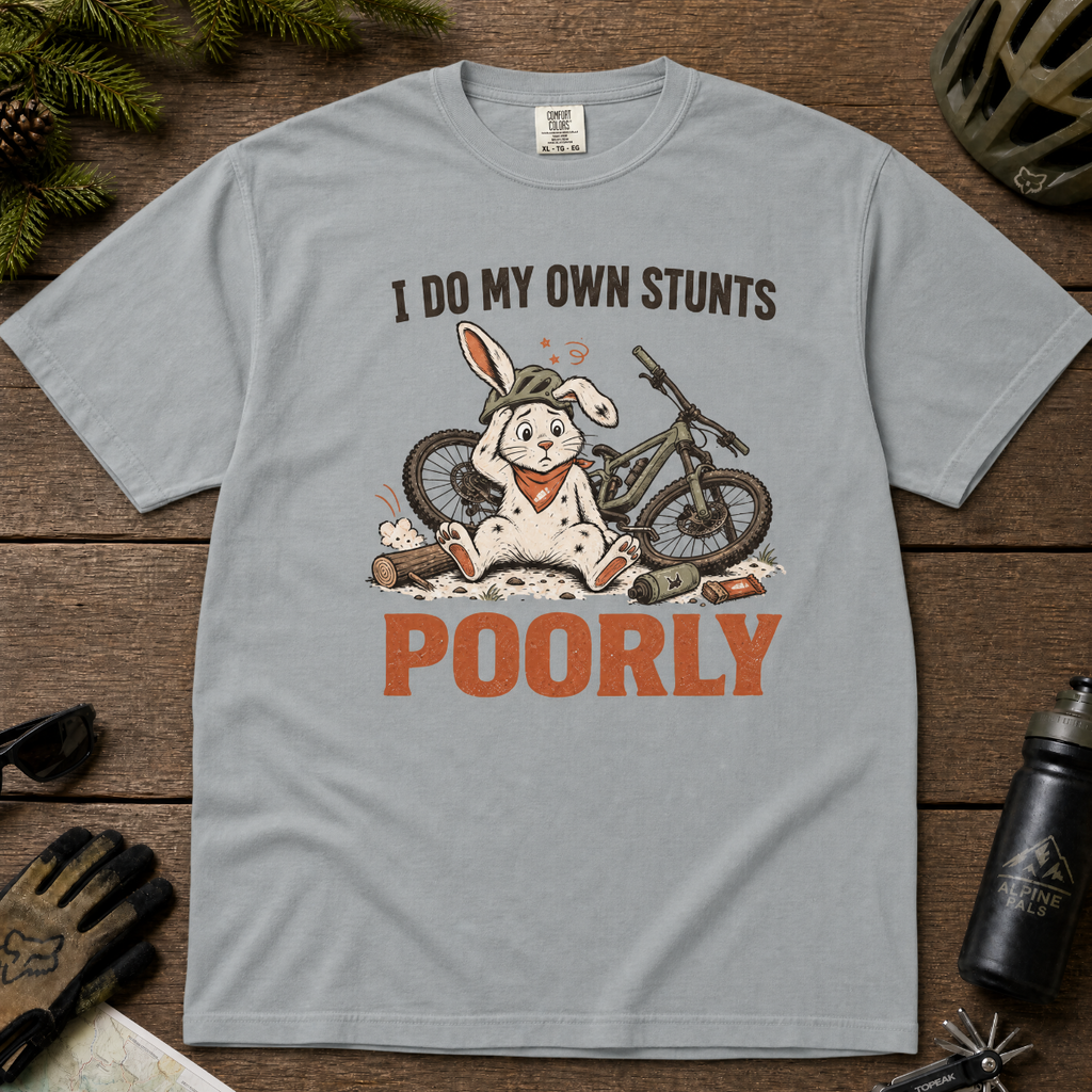 Granite gray t-shirt with a graphic of a rabbit and bicycle, text 'I do my own stunts poorly', on a wooden surface with outdoor gear.
