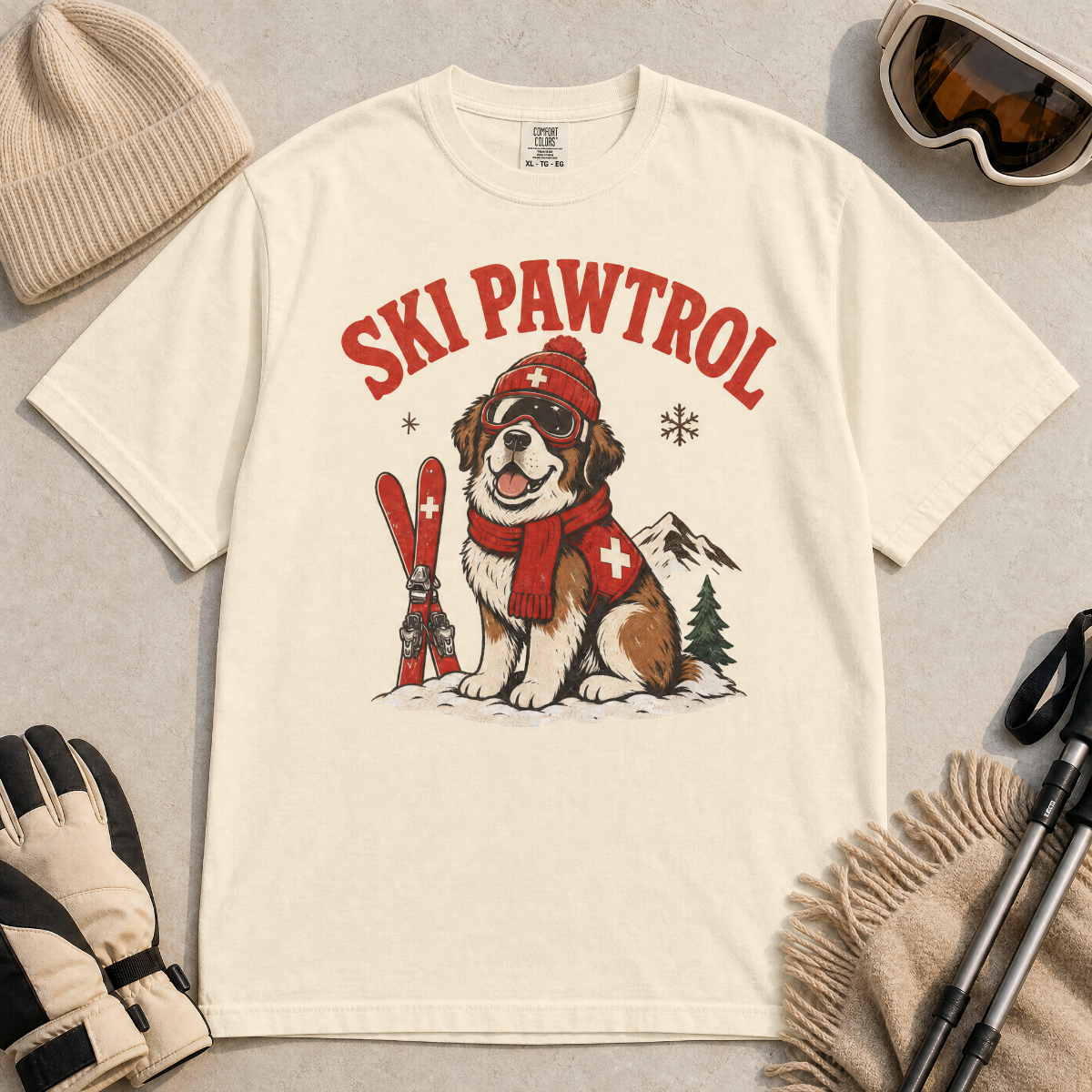 Ivory white t-shirt with a dog graphic and 'Ski Pawtrol' text, surrounded by winter accessories on a light background.