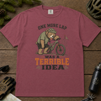 Brick red t-shirt with bear on a bike graphic and text on a wooden surface with cycling accessories.