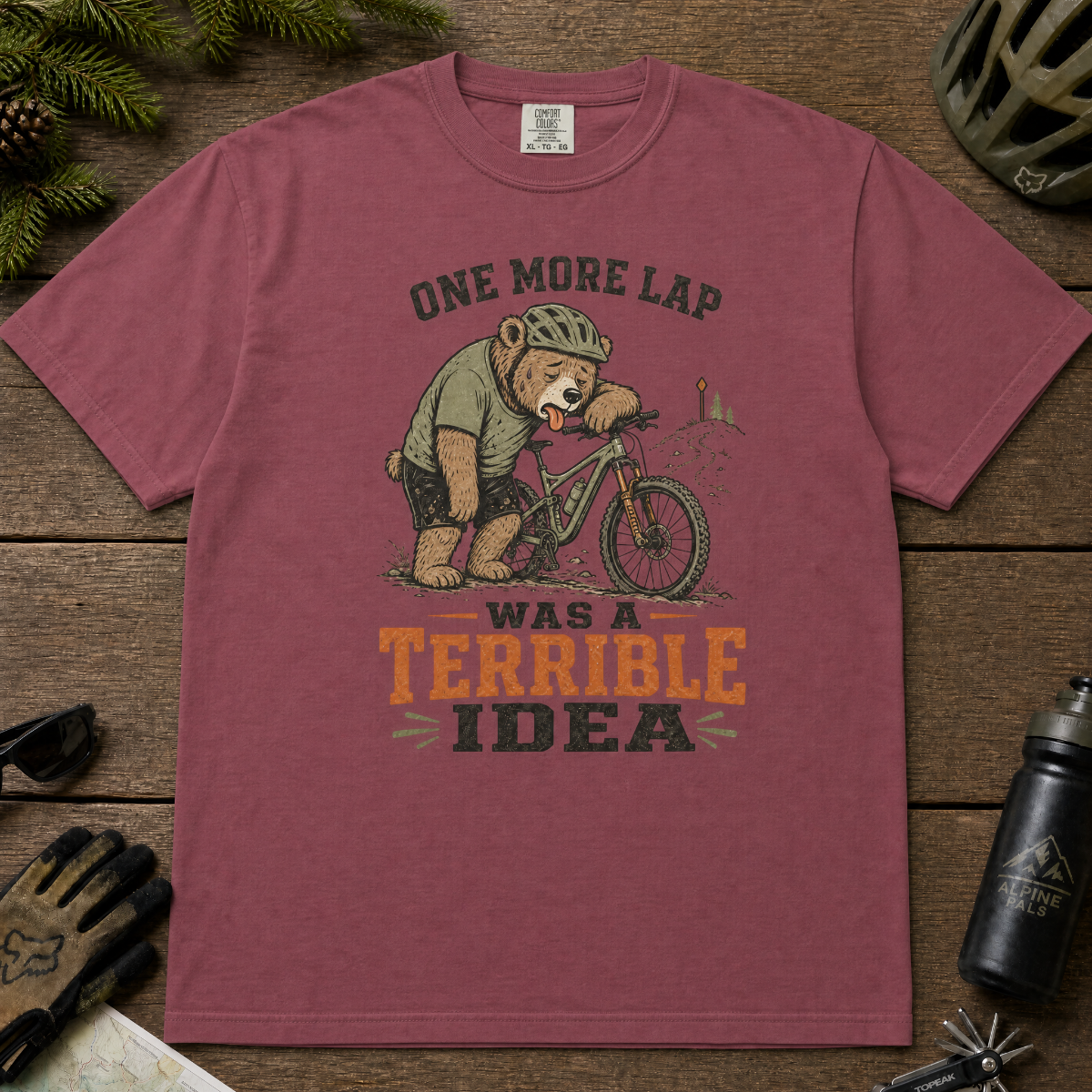 Brick red t-shirt with bear on a bike graphic and text on a wooden surface with cycling accessories.