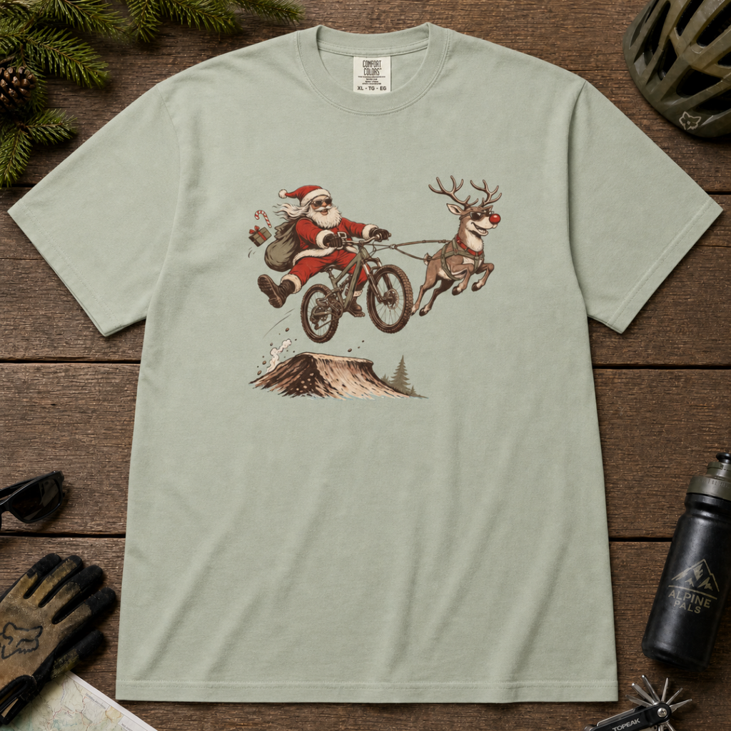 Bay green t-shirt with a graphic of Santa Claus and reindeer on a bike, placed on a wooden surface with outdoor gear.