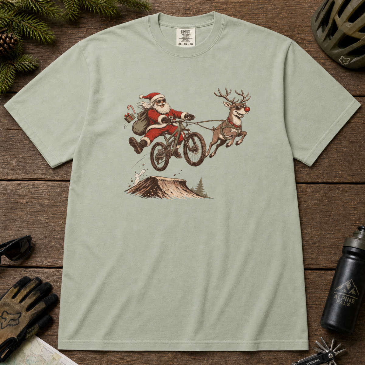 Bay green t-shirt with a graphic of Santa Claus and reindeer on a bike, placed on a wooden surface with outdoor gear.
