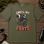 Hemp green t-shirt with a graphic of a skunk on a bike and text 'Smell My Farts' on a wooden surface.