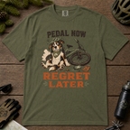 Hemp green t-shirt with a dog and bicycle graphic and 'Pedal Now Regret Later' text on a wooden surface.