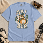 Washed denim blue t-shirt with a dog wearing a hat and goggles graphic, surrounded by skiing accessories on a beige background.
