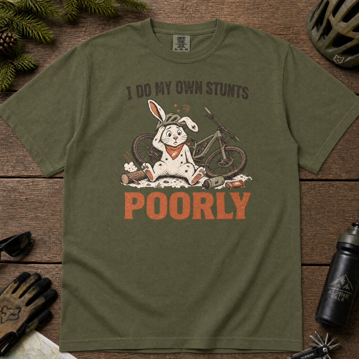 Hemp green t-shirt with a graphic of a rabbit and text 'I do my own stunts poorly' on a wooden surface.