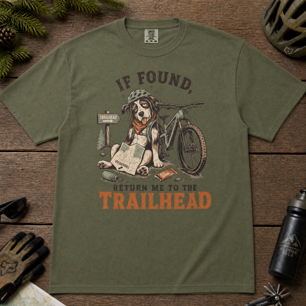 Hemp green t-shirt with a dog graphic and text 'If found, return me to the trailhead' on a wooden surface with outdoor gear.