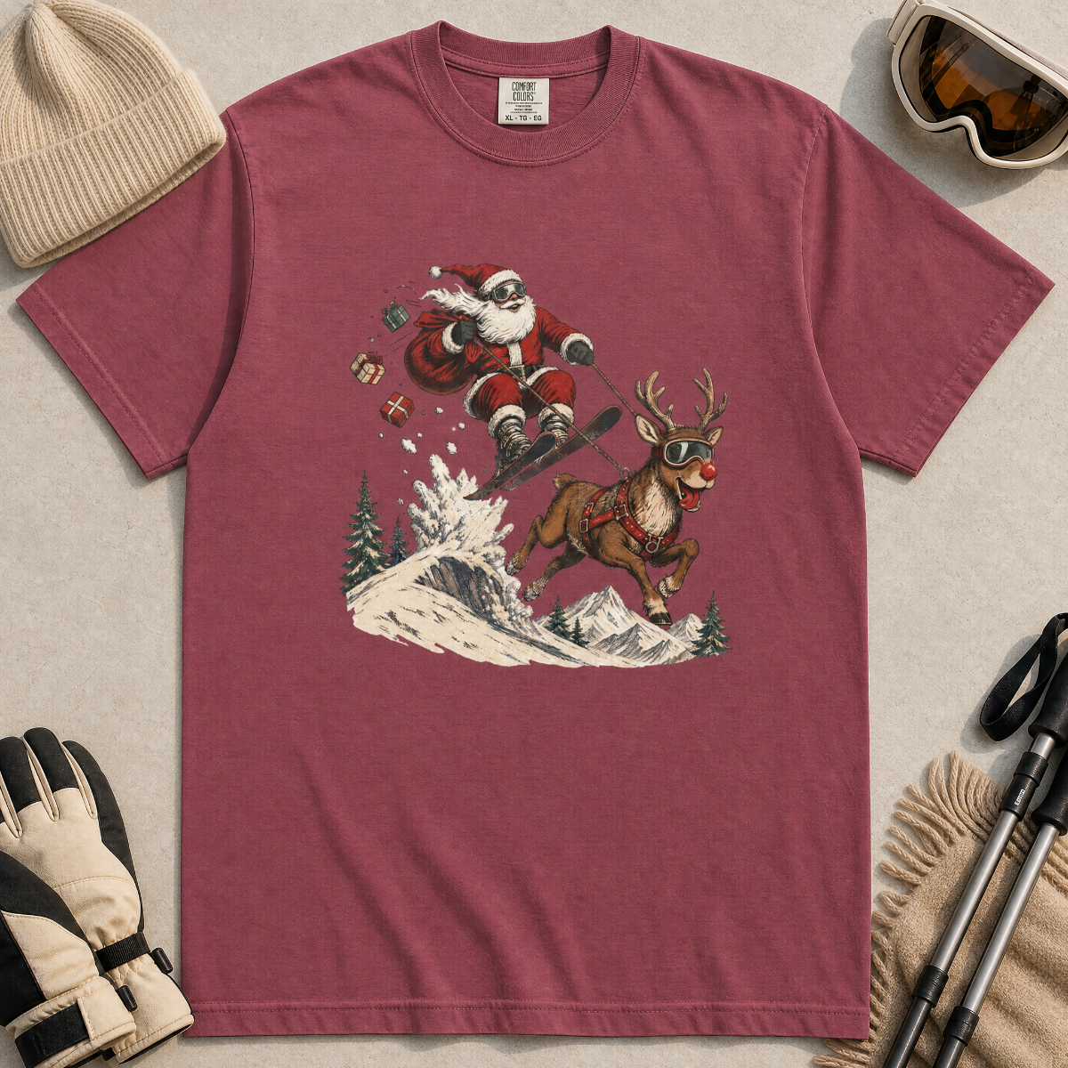 Brick red t-shirt with Santa Claus and reindeer graphic, surrounded by winter accessories on a light background.