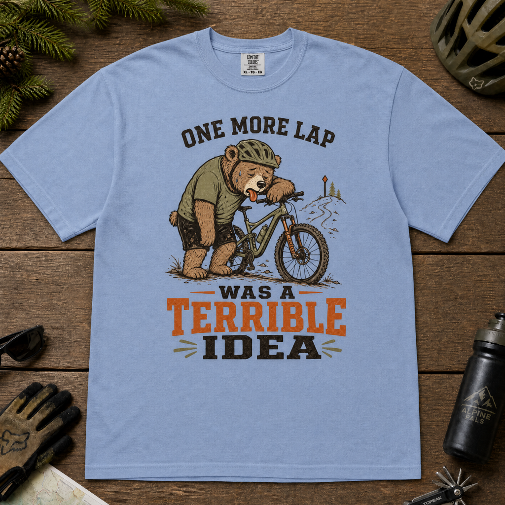 Washed denim blue t-shirt with a bear on a bicycle graphic and text 'One more lap was a terrible idea' on a wooden surface.