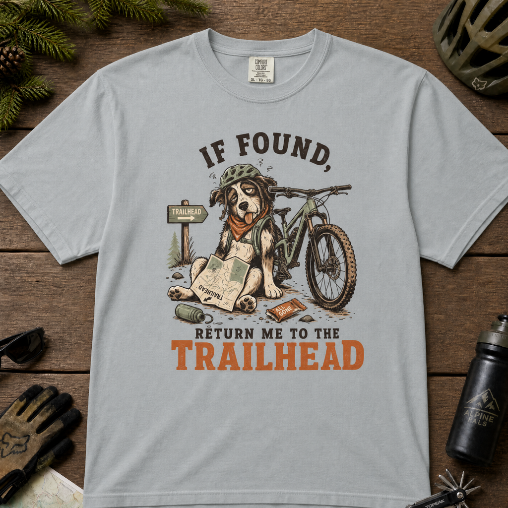 Granite gray t-shirt with a graphic of a dog with a bicycle and text 'If Found, Return Me to the Trailhead' on a wooden surface with outdoor gear.