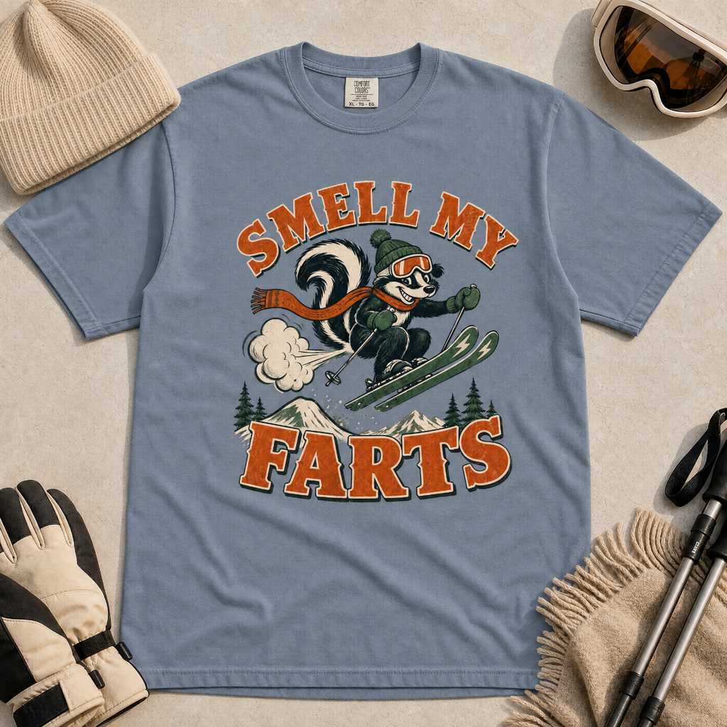 Blue jean colored t-shirt with a skunk graphic and text 'Smell my Farts' on a beige surface with winter accessories.