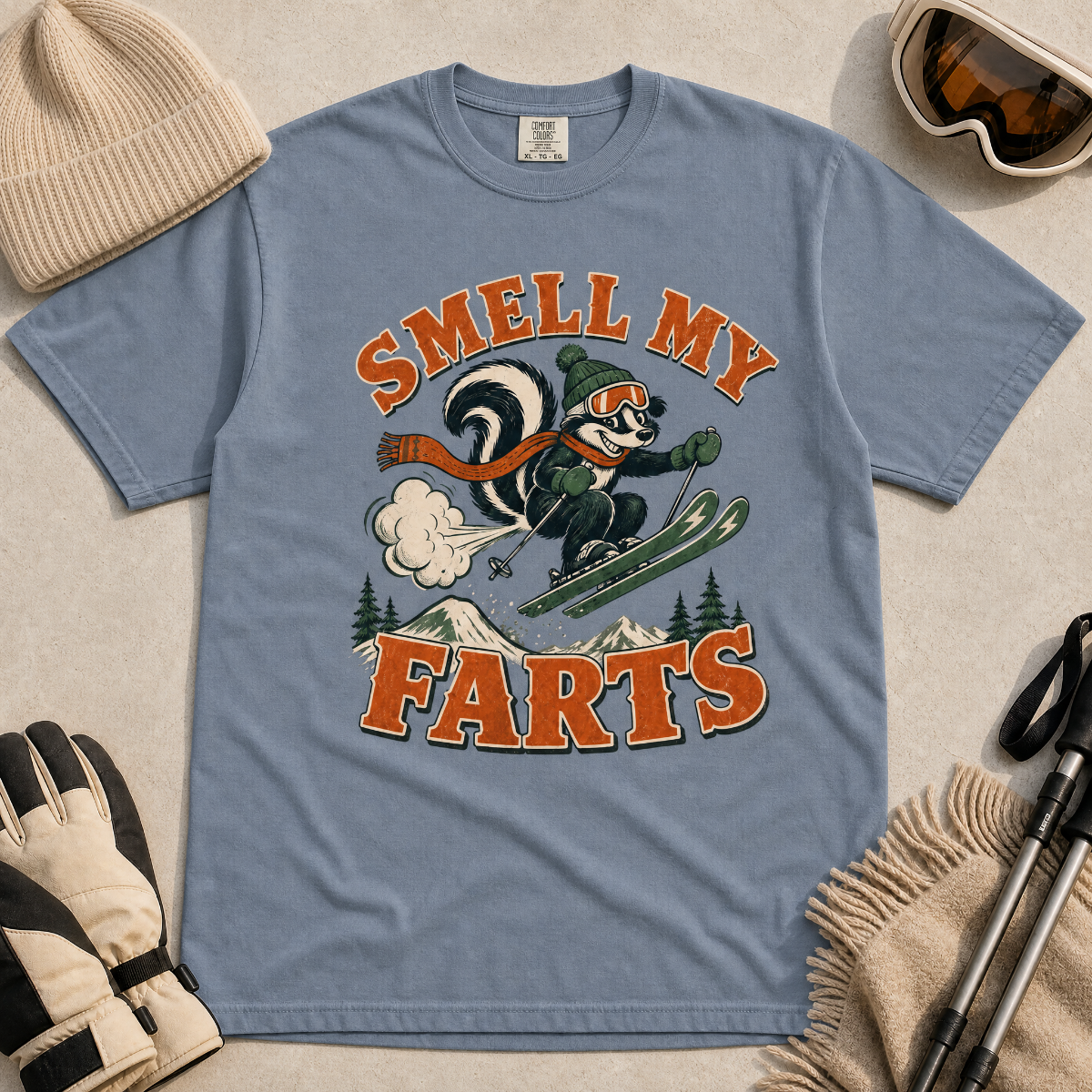 Blue jean colored t-shirt with a skunk graphic and text 'Smell my Farts' on a beige surface with winter accessories.