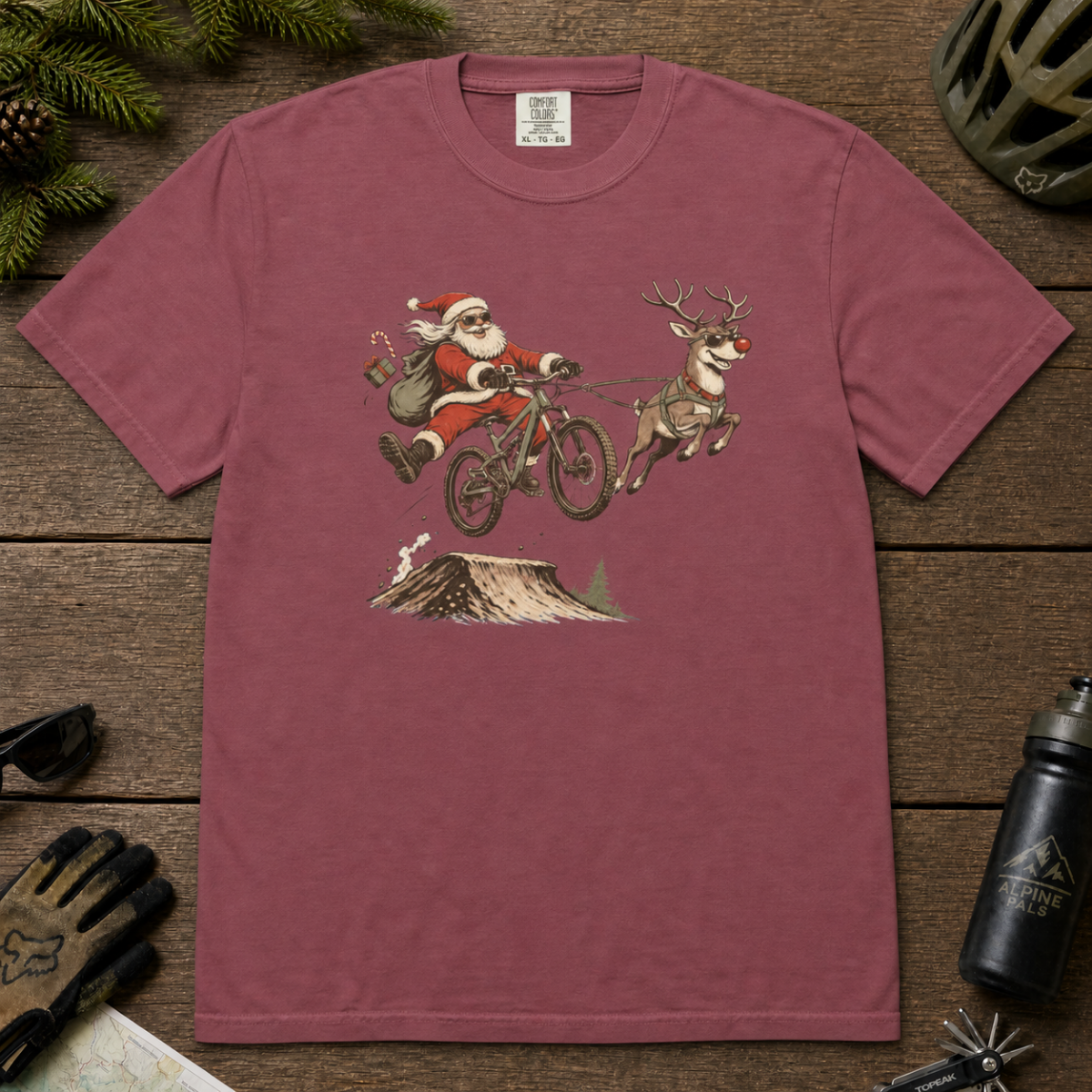 Brick red t-shirt with Santa Claus riding a bike and reindeer design on a wooden surface.