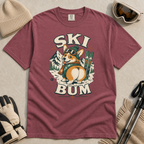 Brick red t-shirt with a dog wearing skis and goggles graphic and 'SKI BUM' text, surrounded by winter accessories on a beige surface.