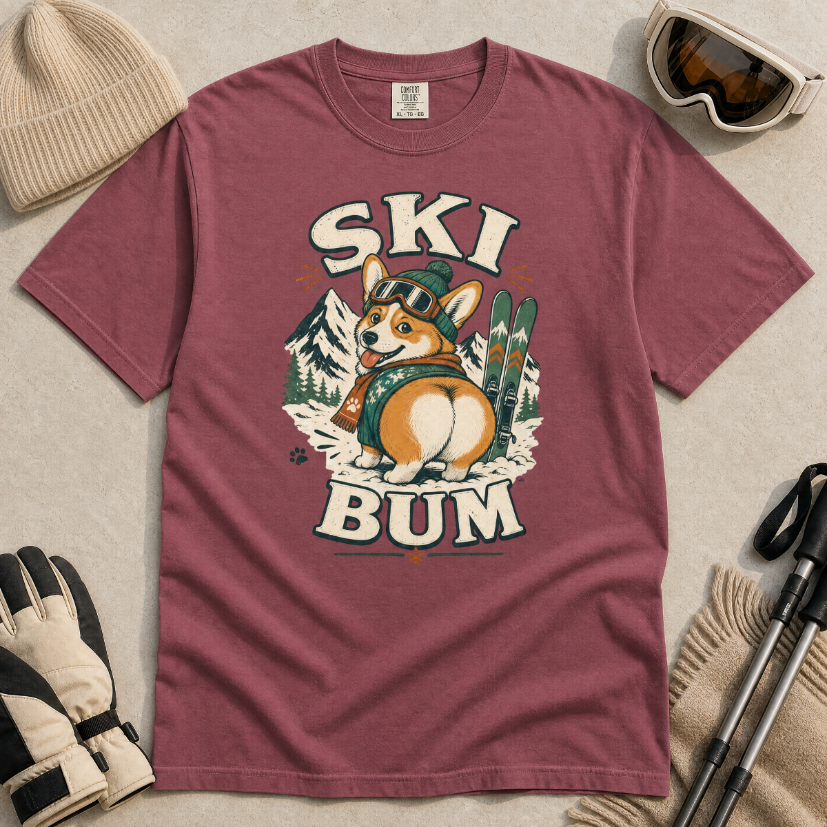 Brick red t-shirt with a dog wearing skis and goggles graphic and 'SKI BUM' text, surrounded by winter accessories on a beige surface.