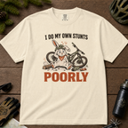 Ivory white t-shirt with a graphic of a rabbit on a bike and text 'I do my own stunts poorly' on a wooden surface.