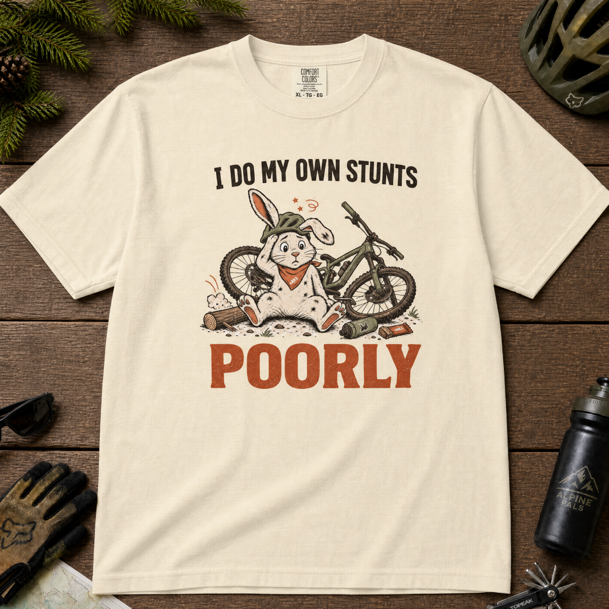 Ivory white t-shirt with a graphic of a rabbit on a bike and text 'I do my own stunts poorly' on a wooden surface.