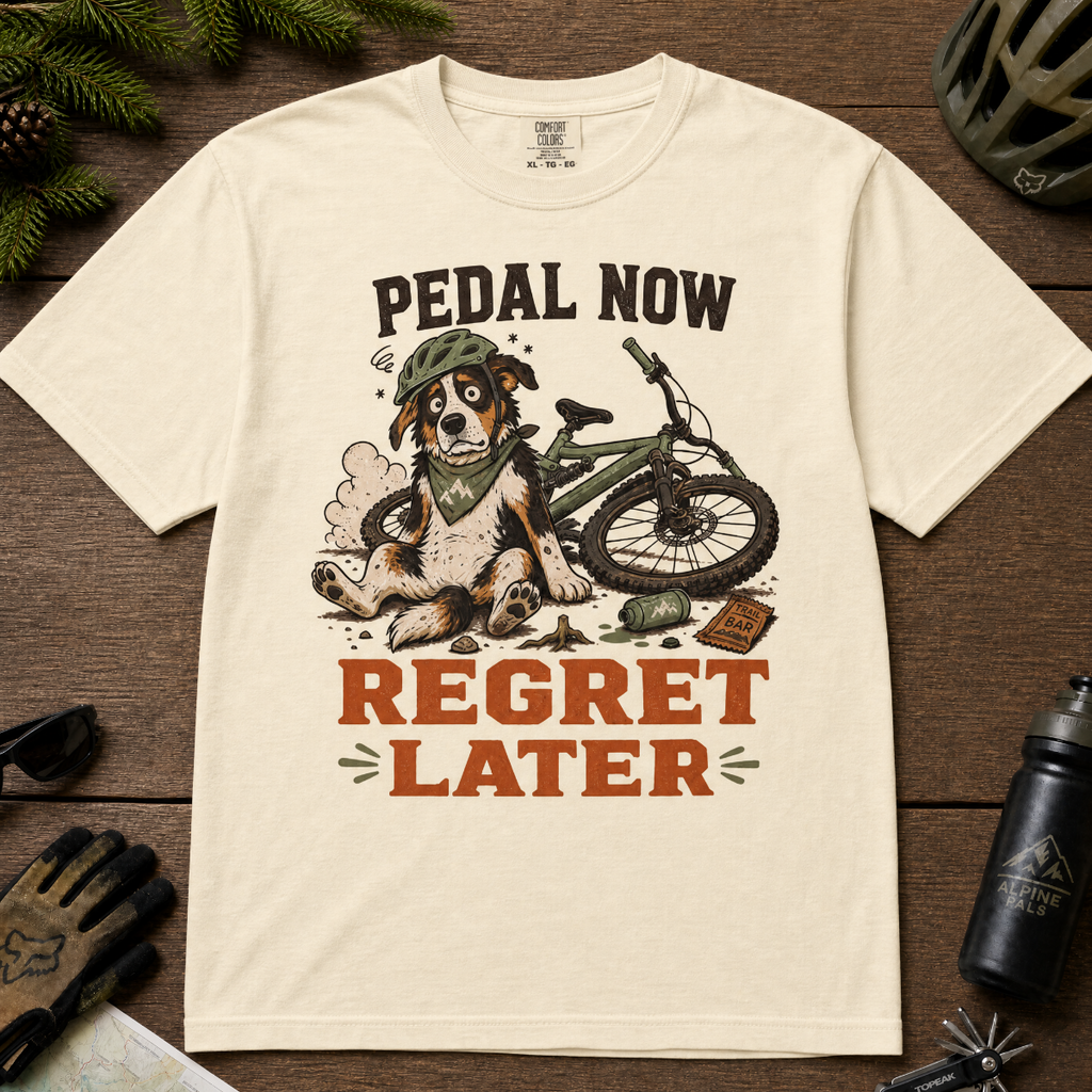 Ivory white t-shirt with a graphic of a dog with a bike and text 'Pedal Now Regret Later' on a wooden surface.