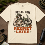 Ivory white t-shirt with a graphic of a dog with a bike and text 'Pedal Now Regret Later' on a wooden surface.