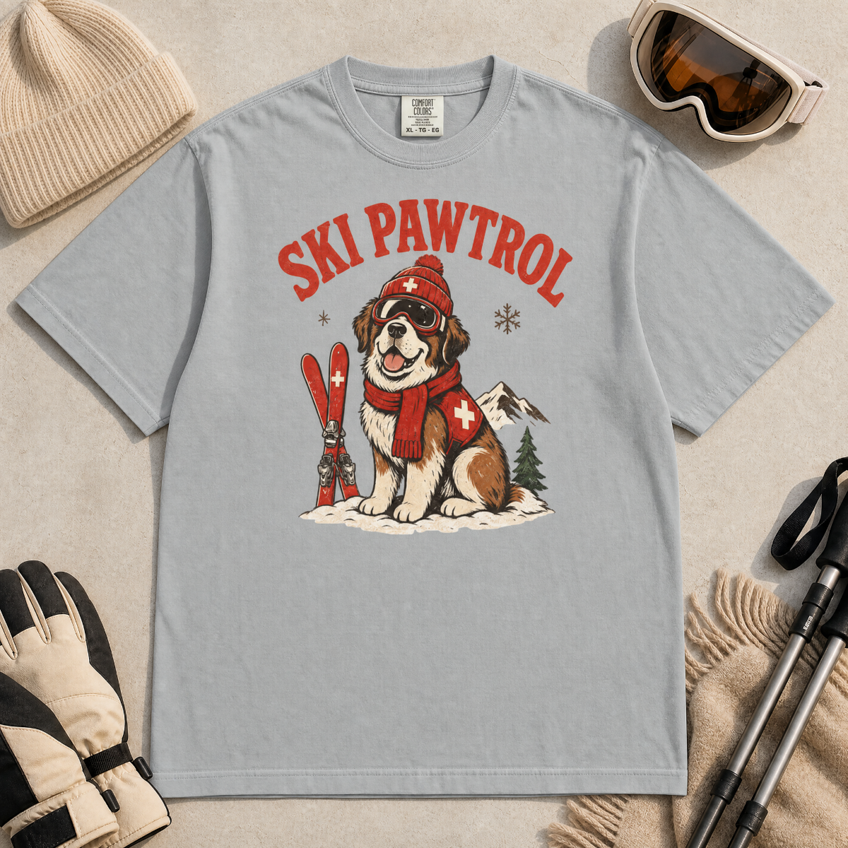 Granite gray t-shirt with a dog graphic and 'Ski Pawtrol' text, surrounded by winter accessories on a beige background.