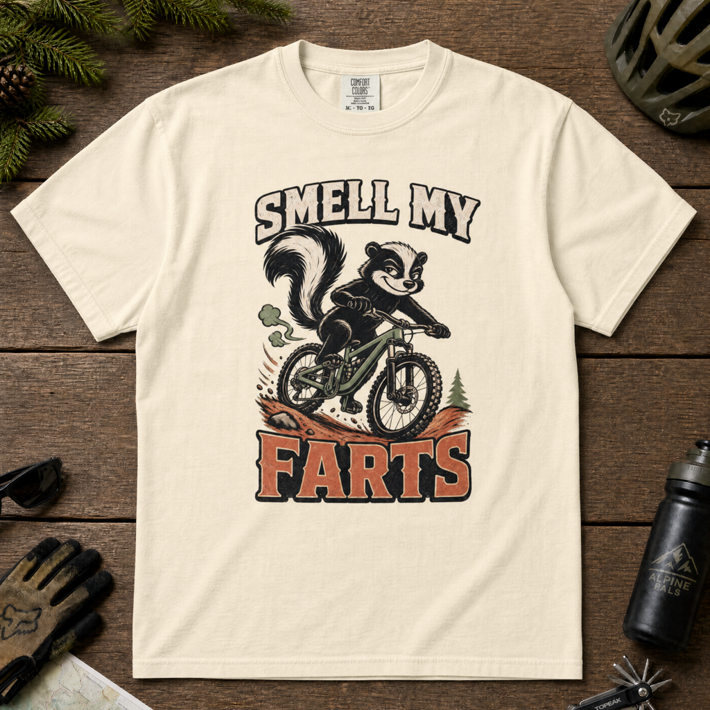 Ivory white t-shirt with a graphic of a skunk on a bike and text 'SMELL MY FARTS' on a wooden surface.