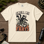 Ivory white t-shirt with a graphic of a skunk on a bike and text 'SMELL MY FARTS' on a wooden surface.